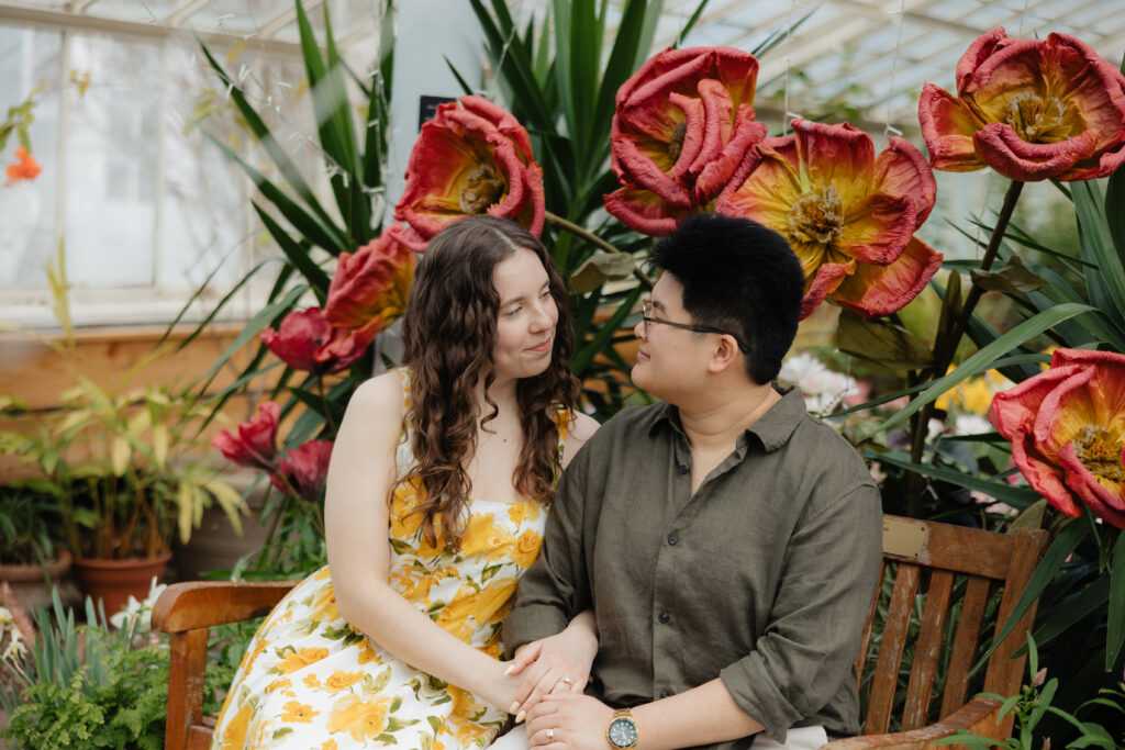 Couple sitting inside a bright greenhouse surrounded by spring flowers at Buffalo Botanical Gardens engagement shoot