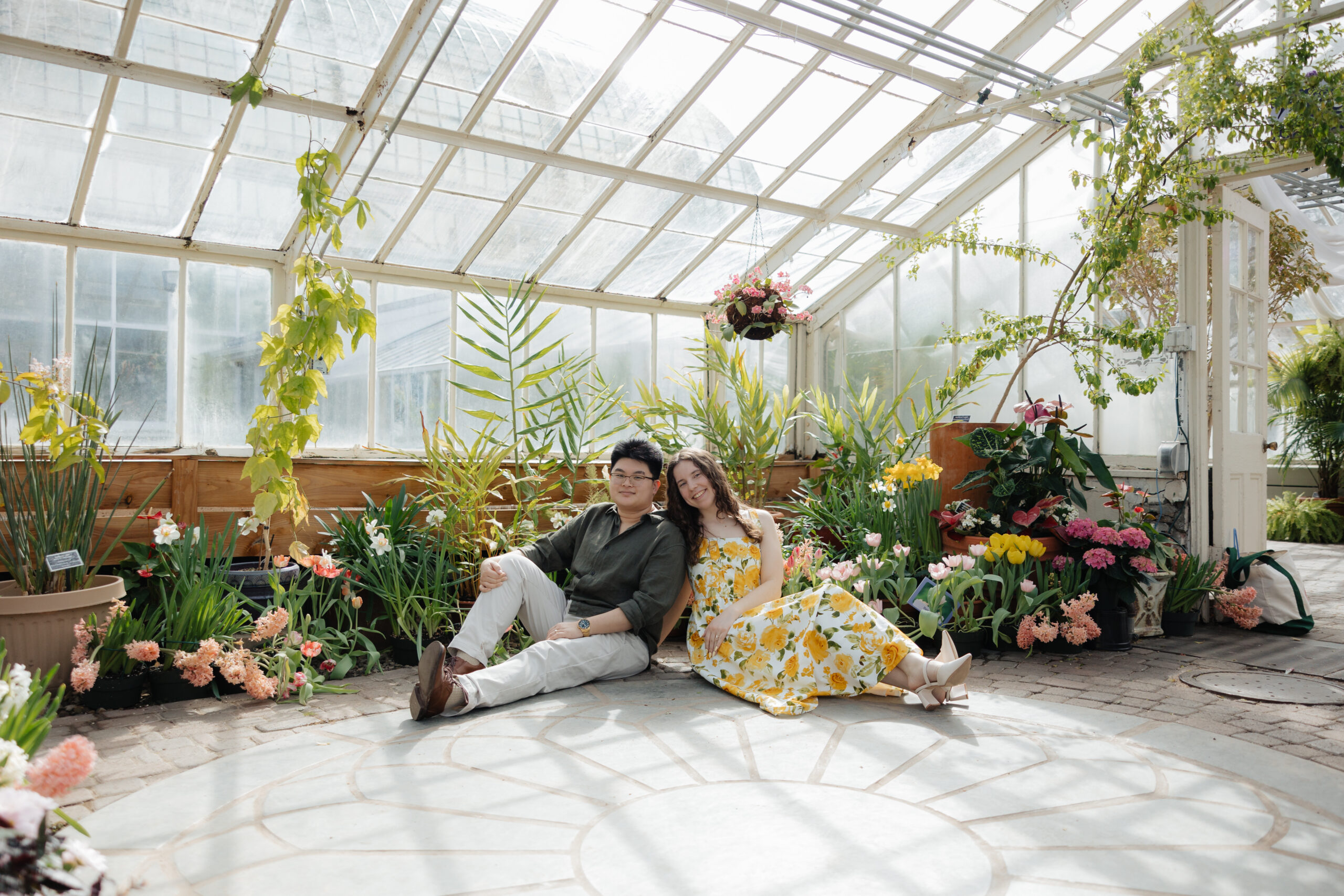 Couple sitting together inside Buffalo and Erie County Botanical Gardens greenhouse during spring engagement session surrounded by flowers and natural light.